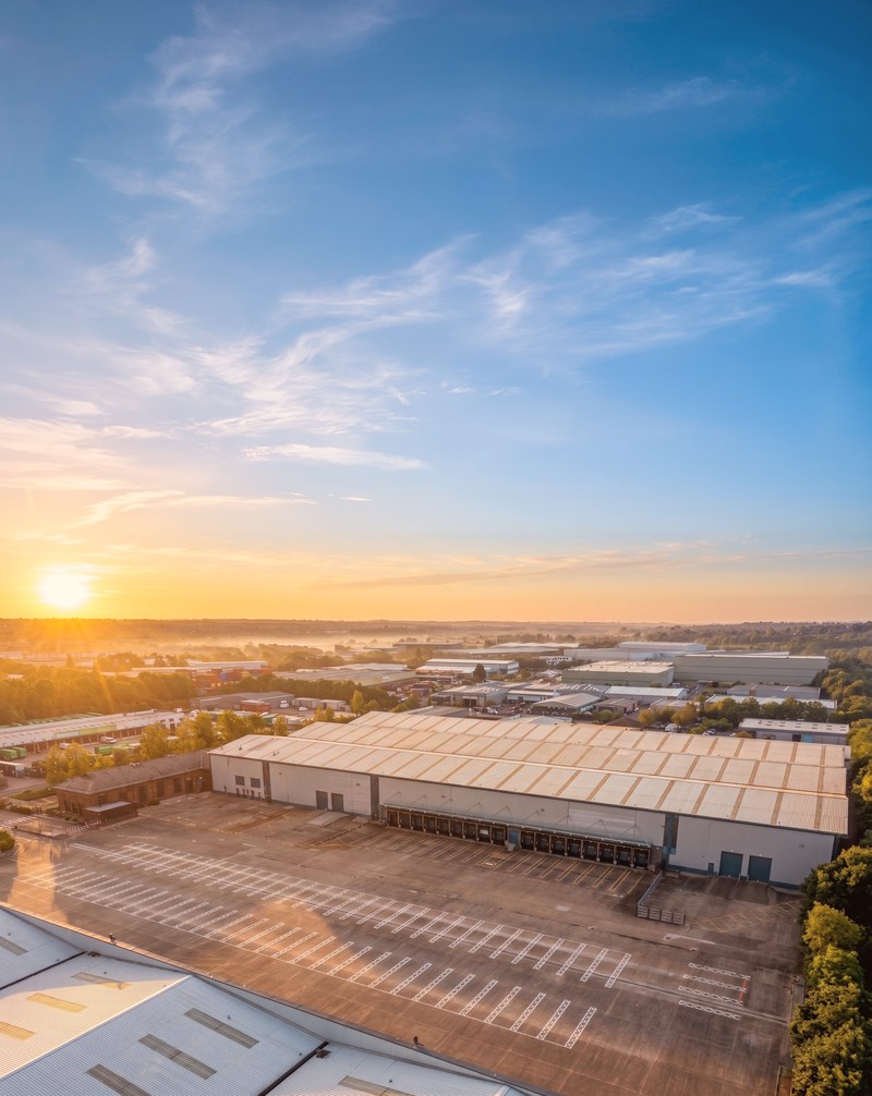 Brackmills 195 from the air at sunrise: a long warehouse with a row of dock doors behind a wide, empty marked yard, the Brackmills estate and morning mist beyond